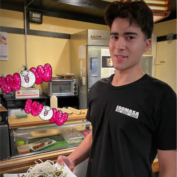 A young Edomasa Sushi Bar & Restaurant employee holds a plate of food in a kitchen.