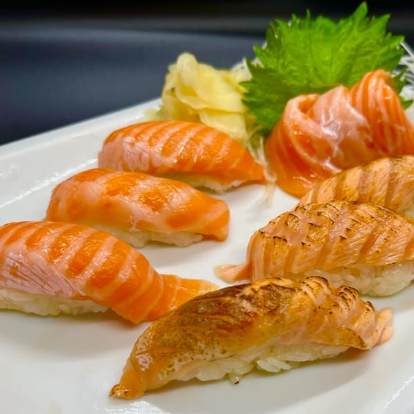 A white plate holds various salmon sushi and sashimi, garnished with green leaves and pickled ginger.